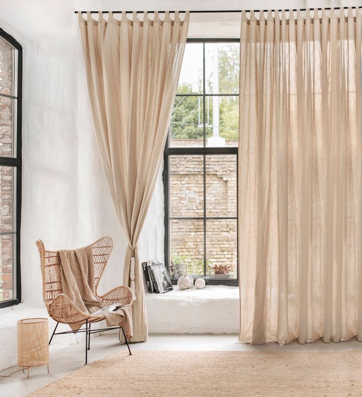 Beige sheer curtains on black-framed windows in a cozy, minimalist living room with a rattan chair.