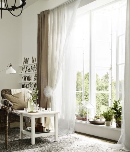 Bright living room with sheer curtains, large windows, indoor plants, and cozy wicker furniture