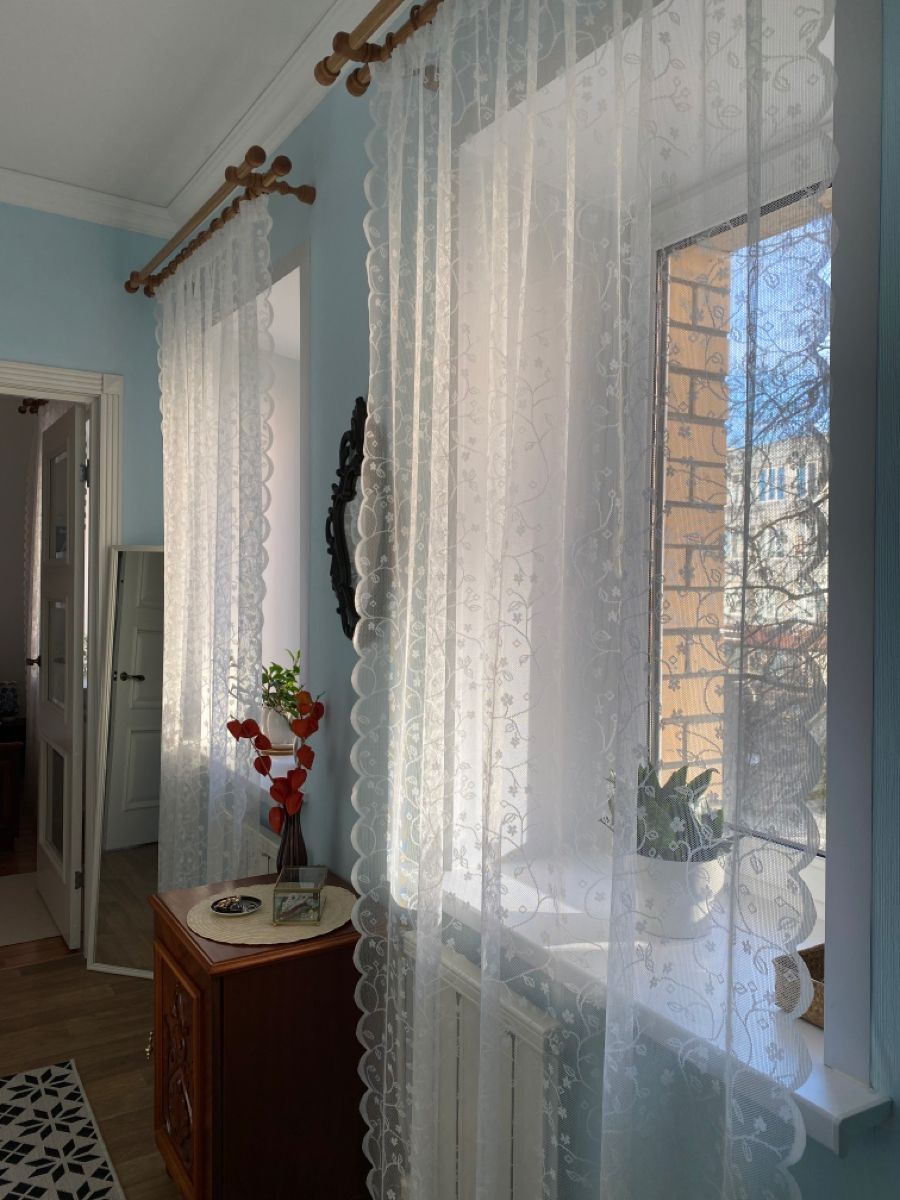 Sheer white lace curtains on wooden rods in a sunlit, cozy living room with light blue walls.