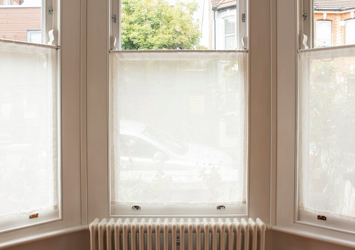 Bright bay window with sheer white curtain panels and a radiator below.