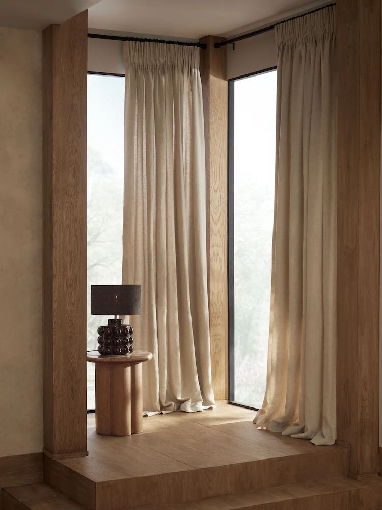Beige blackout curtains on tall windows in a modern, light wood corner with a lamp and side table.