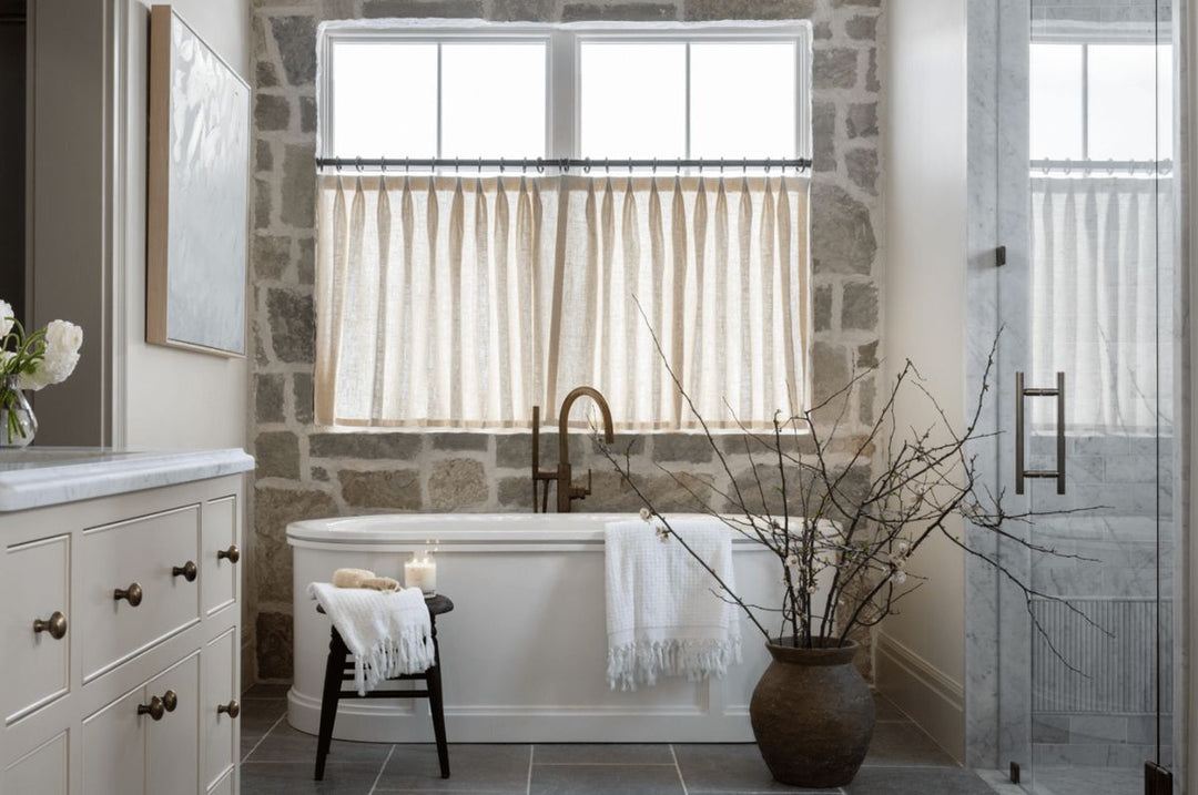 Neutral curtain panels on windows in a modern stone-wall bathroom with a freestanding tub