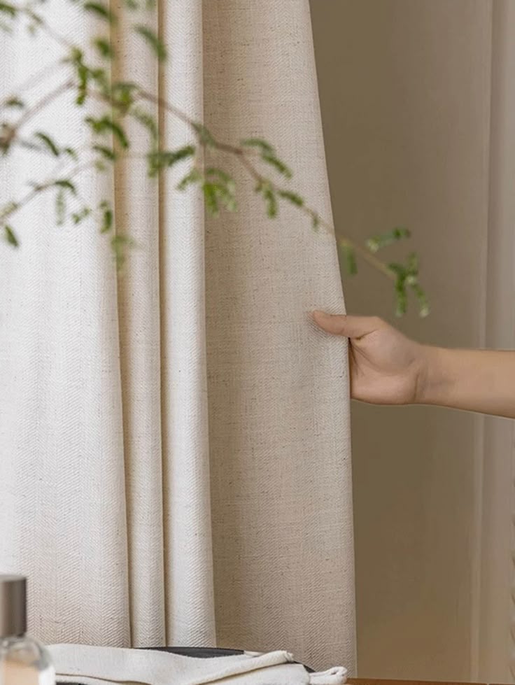 Hand pulling beige blackout curtain with thermal fabric in a cozy home setting