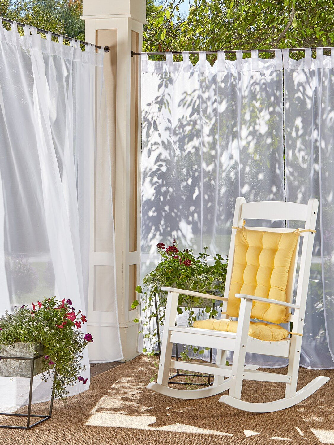Outdoor patio with sheer white curtains, white rocking chair with yellow cushion, and potted flowers
