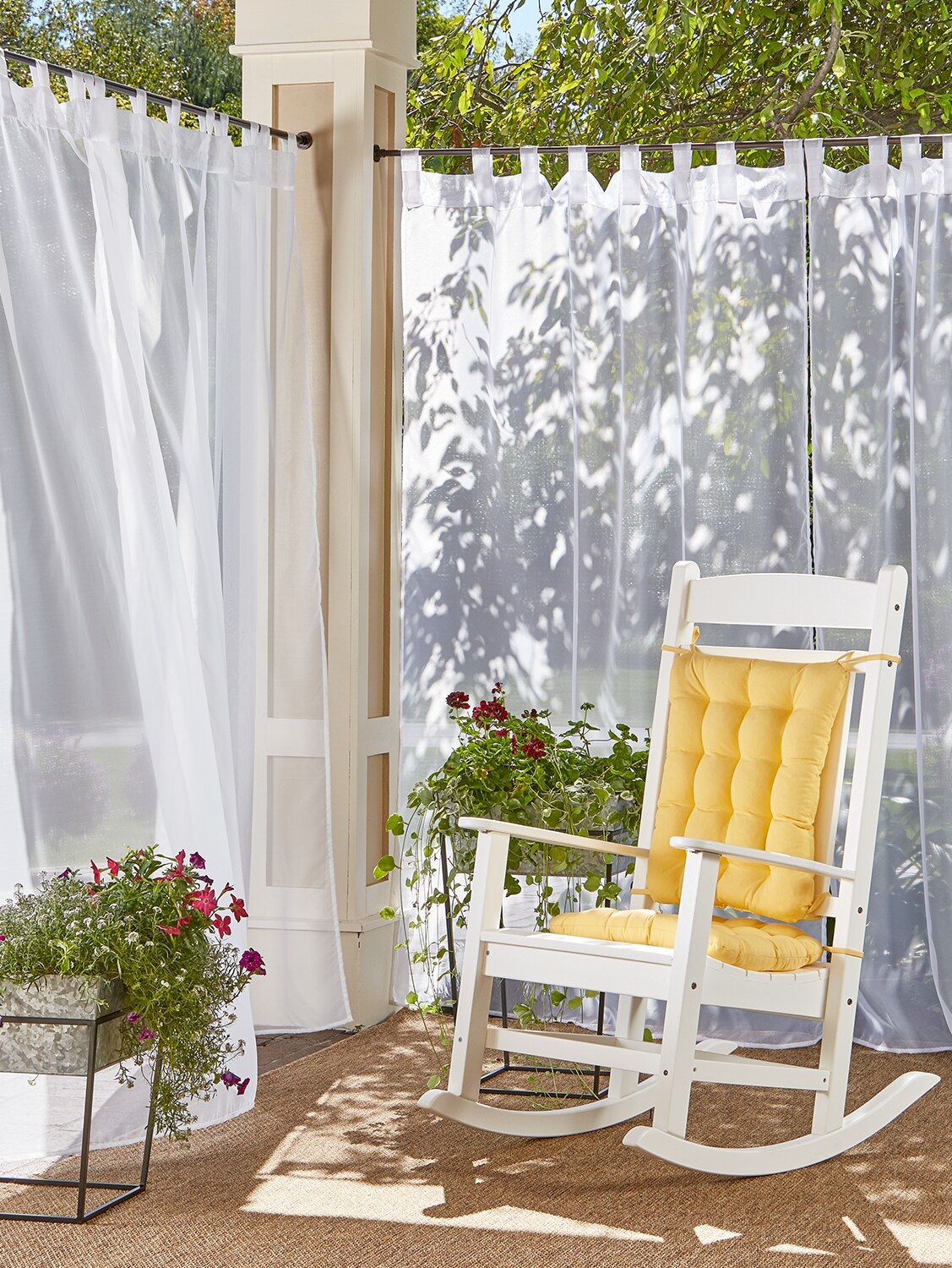 Outdoor patio with sheer white curtains, white rocking chair with yellow cushion, and potted flowers