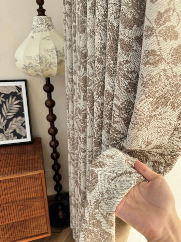 Hand holding beige floral blackout curtain in cozy living room setting