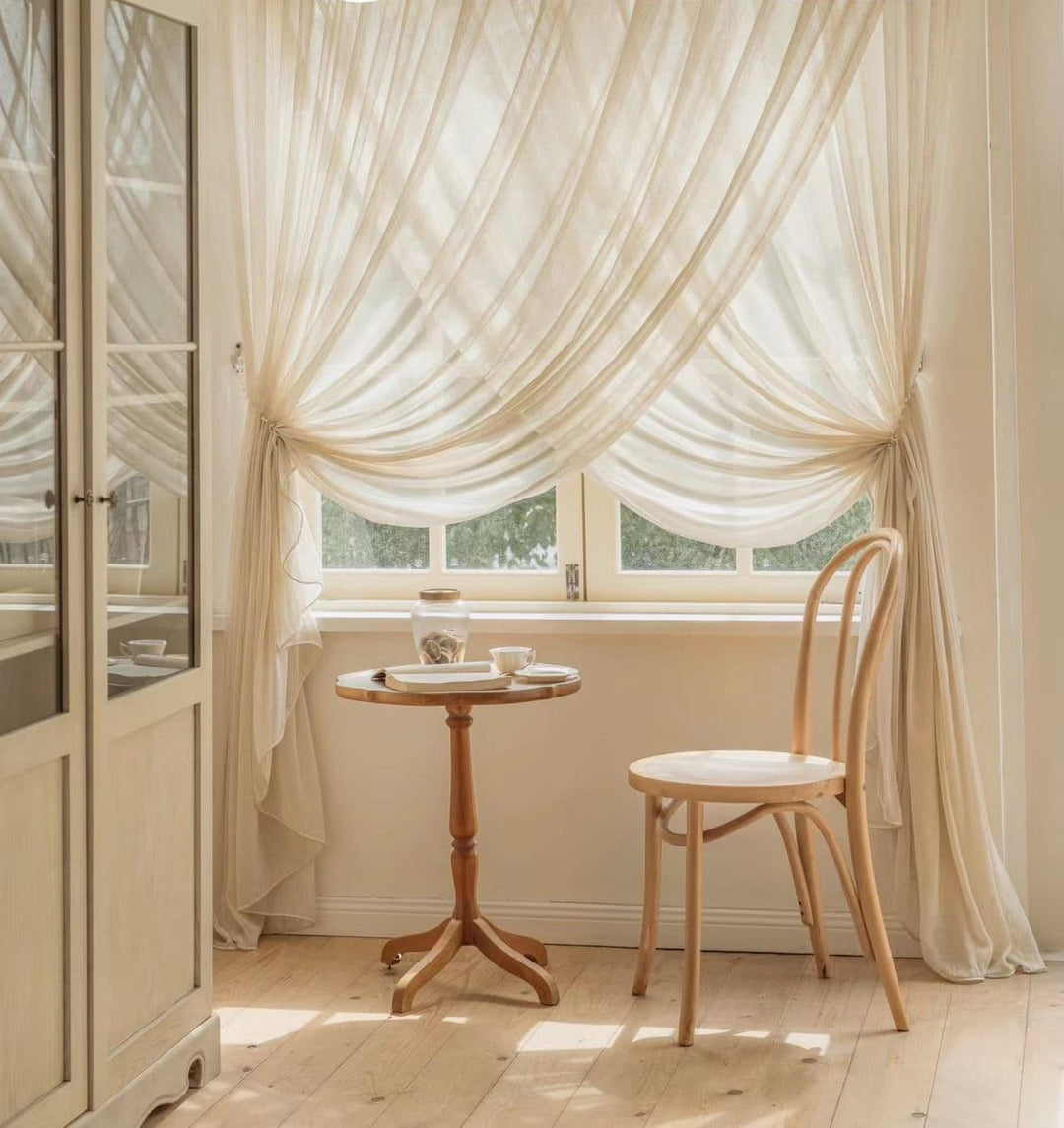 Elegant light beige sheer curtains draped over a bright window in a cozy room with wooden furniture