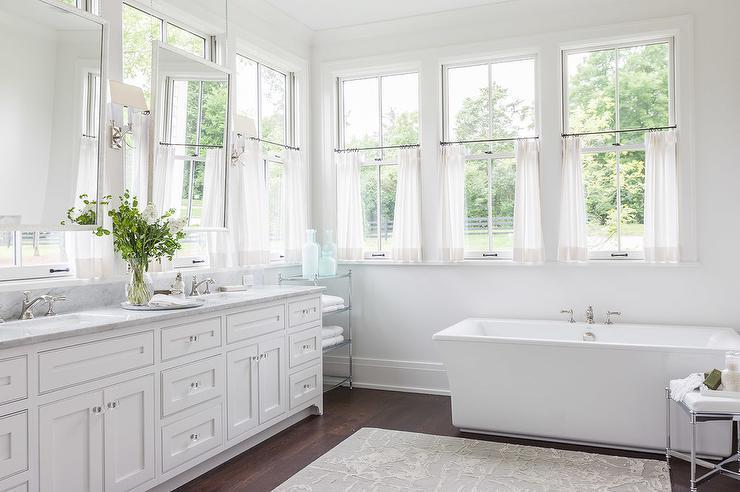 Bright bathroom with white curtain panels, large windows, freestanding tub, and double vanity.