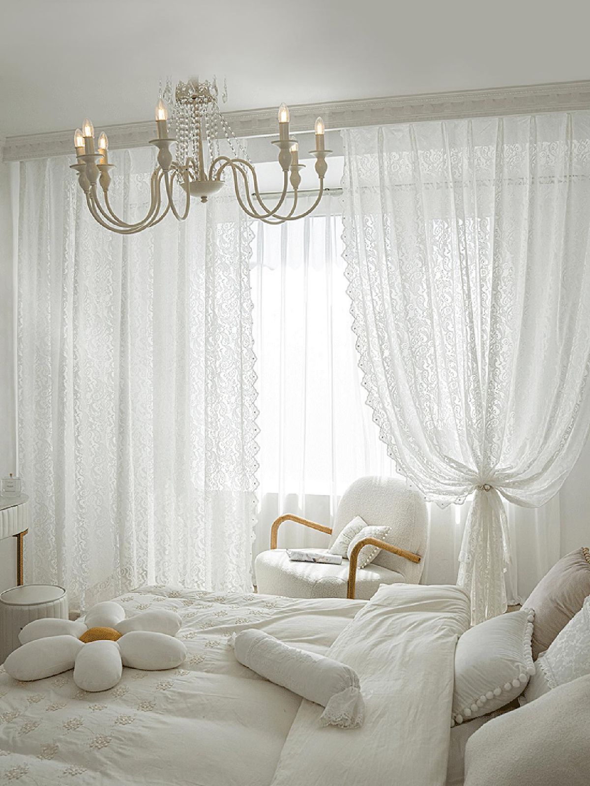 Elegant bedroom with sheer white lace curtains, chandelier, and cozy white bedding.