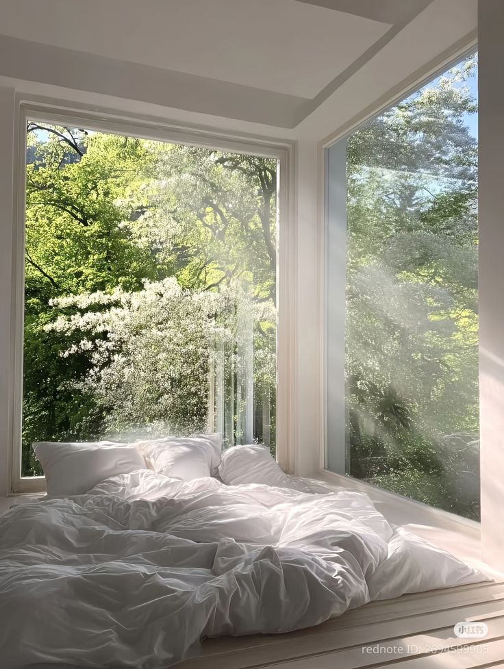 Bright bedroom with white bedding, large corner windows, and lush green trees outside