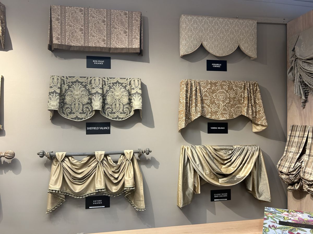 Display of various window valances in different fabrics and styles on a wall