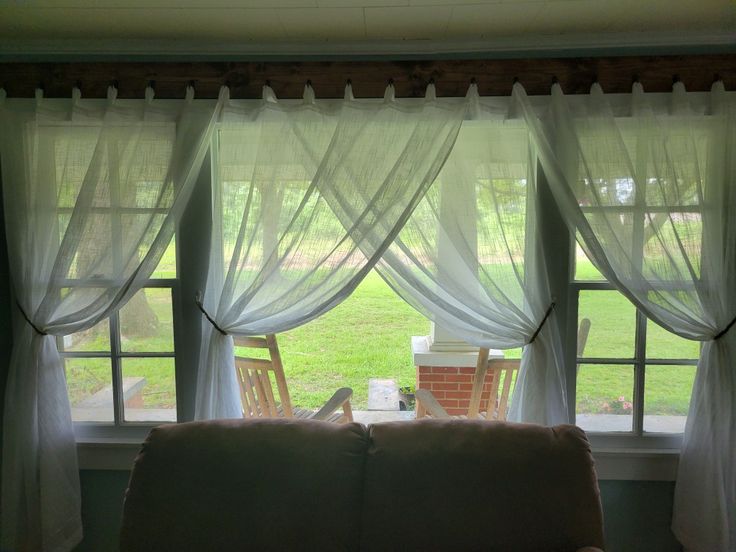 Sheer white curtains with tiebacks on large windows, overlooking a porch with rocking chairs and green lawn