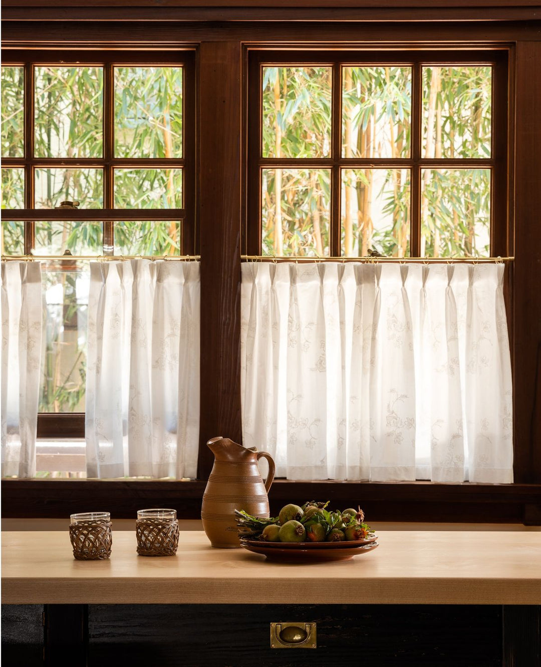Sheer white curtain panels on wooden window, kitchen countertop with ceramic pitcher and fruit plate