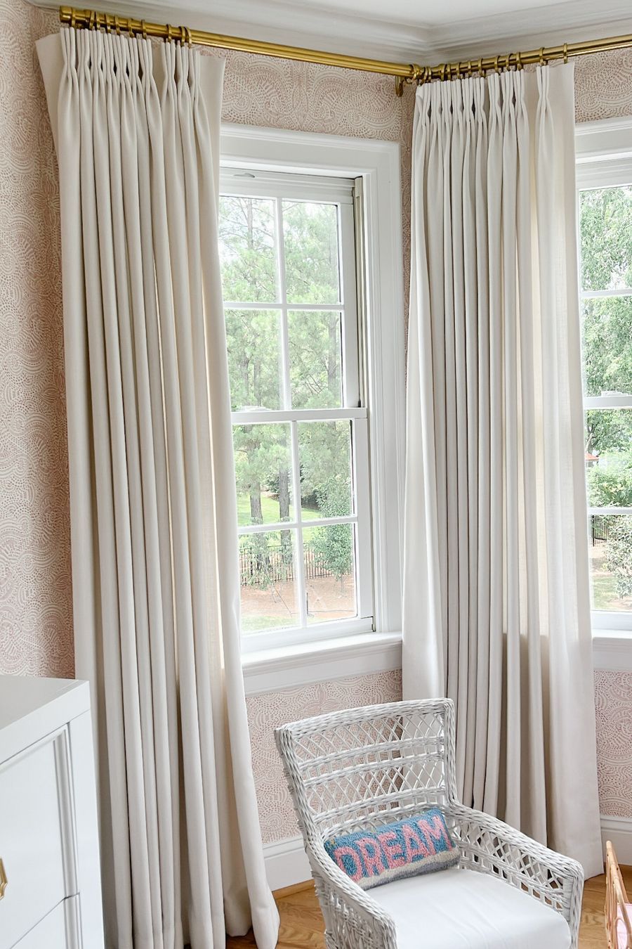 White blackout curtains on brass rod in a bright room with a rattan chair and large window