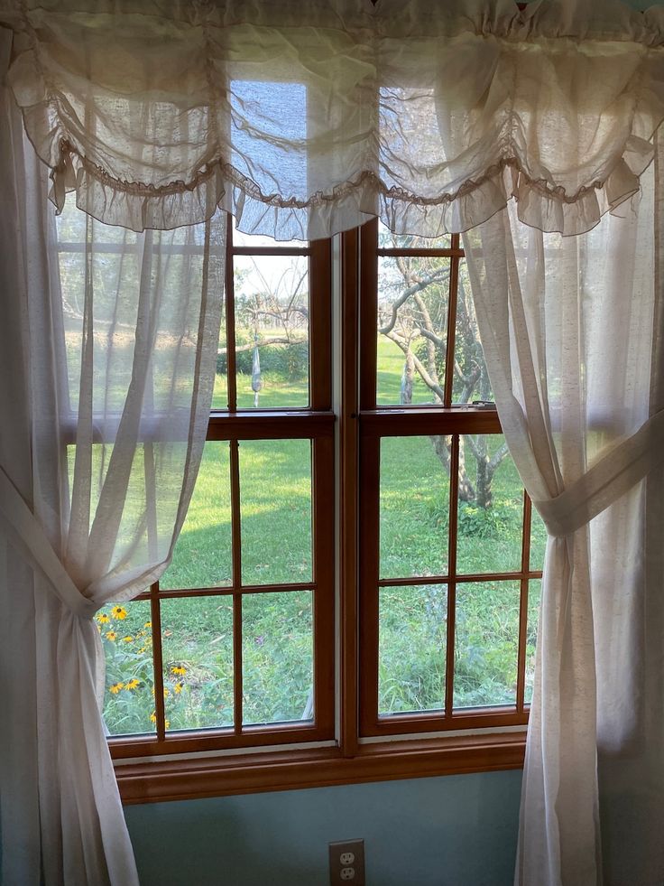 Sheer white curtain panels with ruffled valance on a wooden-framed window, sunlight and garden view