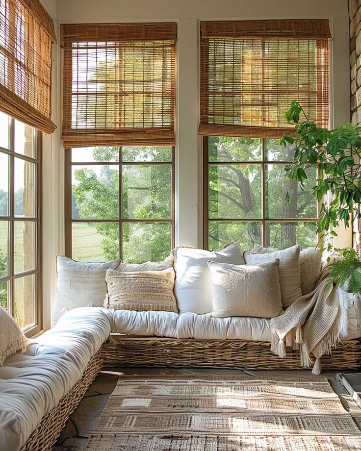 Cozy sunroom with wicker daybed, white pillows, woven shades, and large windows letting in sunlight.