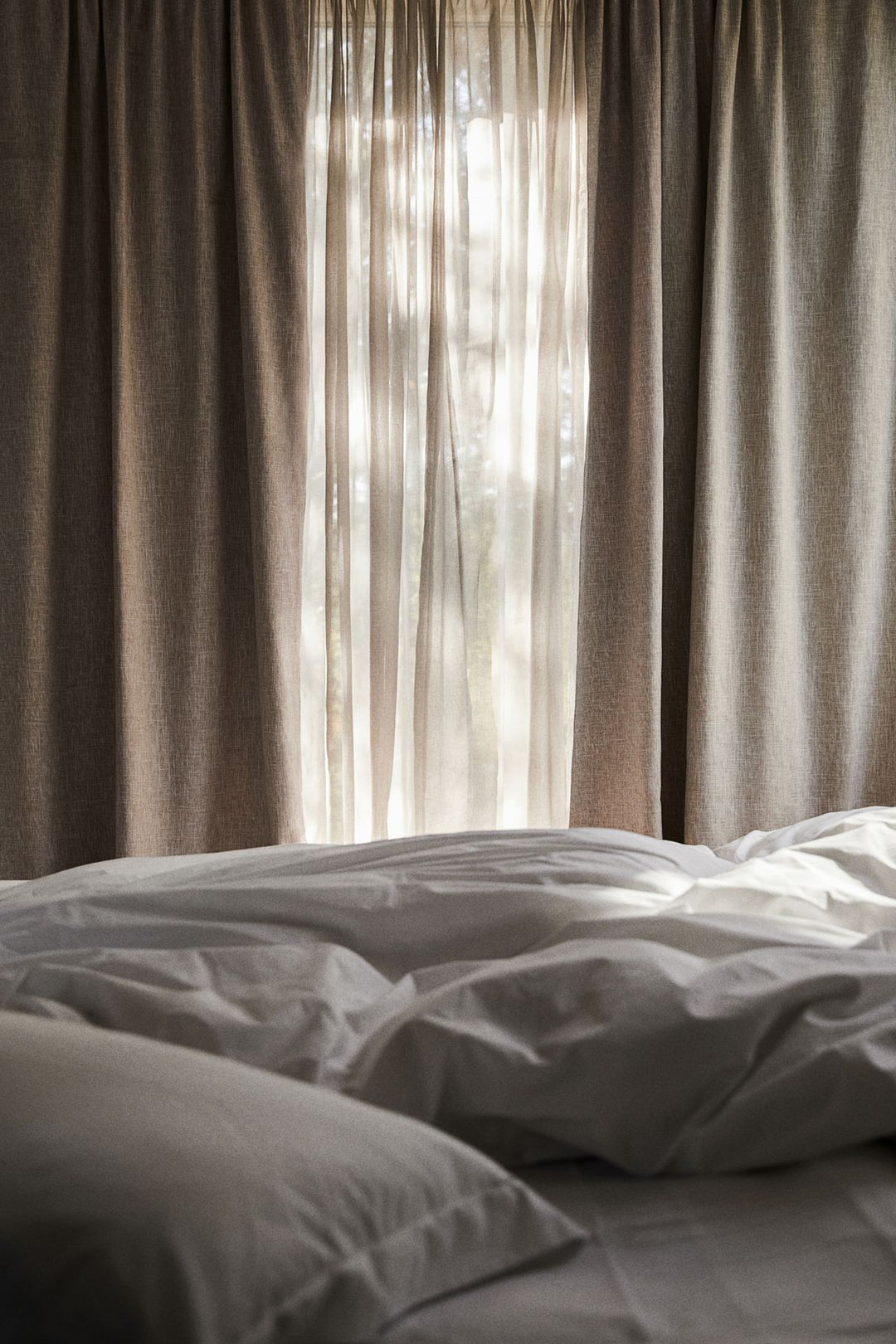 Bedroom window with beige blackout curtains, sheer drapes, and unmade white bedding