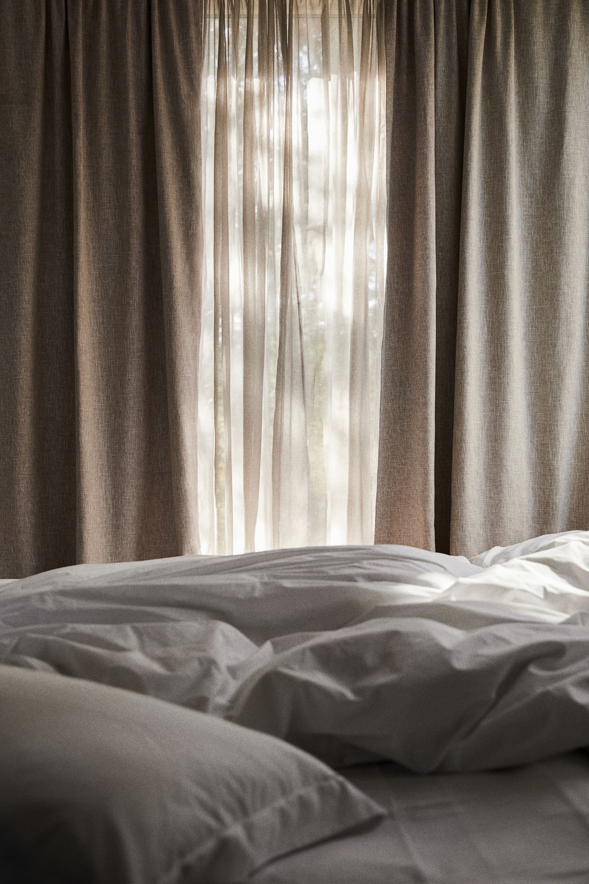 Bedroom window with beige blackout curtains, sheer drapes, and unmade white bedding