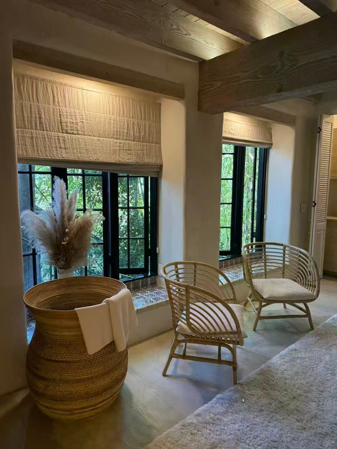 Cozy living space with bamboo chairs, woven basket, Roman shades, and black framed windows.