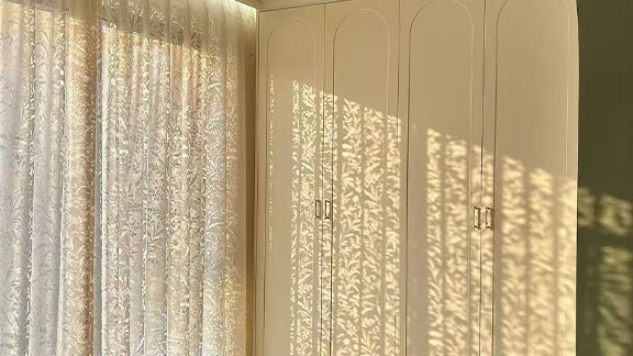 Sheer decorative curtains filtering sunlight, casting patterned shadows on white wardrobe