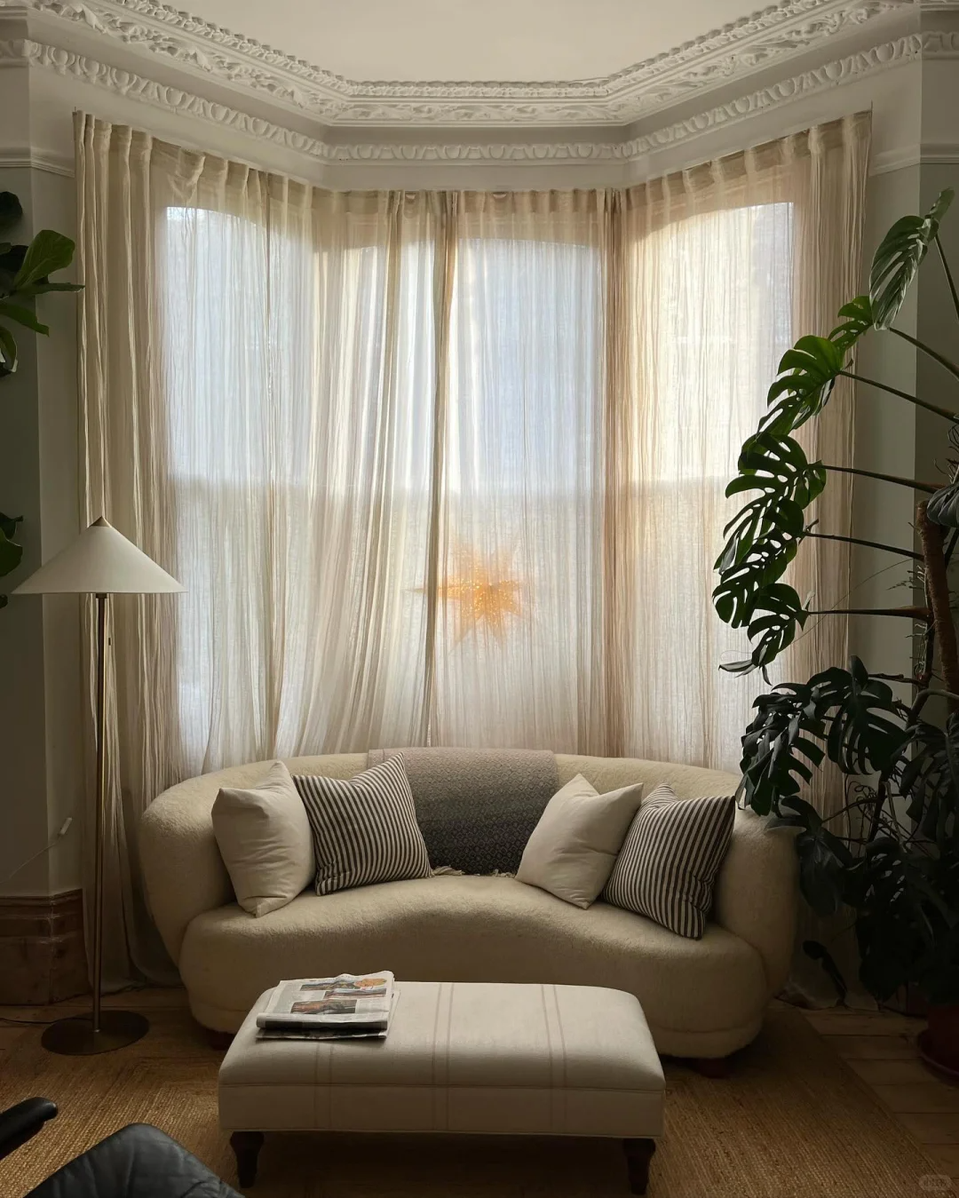 Sheer curtains on bay windows in a cozy living room with cream sofa, striped pillows, and indoor plants.